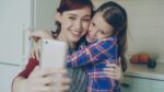 Smiling mother together with funny playful daughter making selfie photo with smartphone camera at home in kitchen. Family, cooking, and people concept